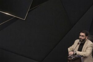 Man med glasögon och skägg i kavaj, bakgrund med geometriska former.